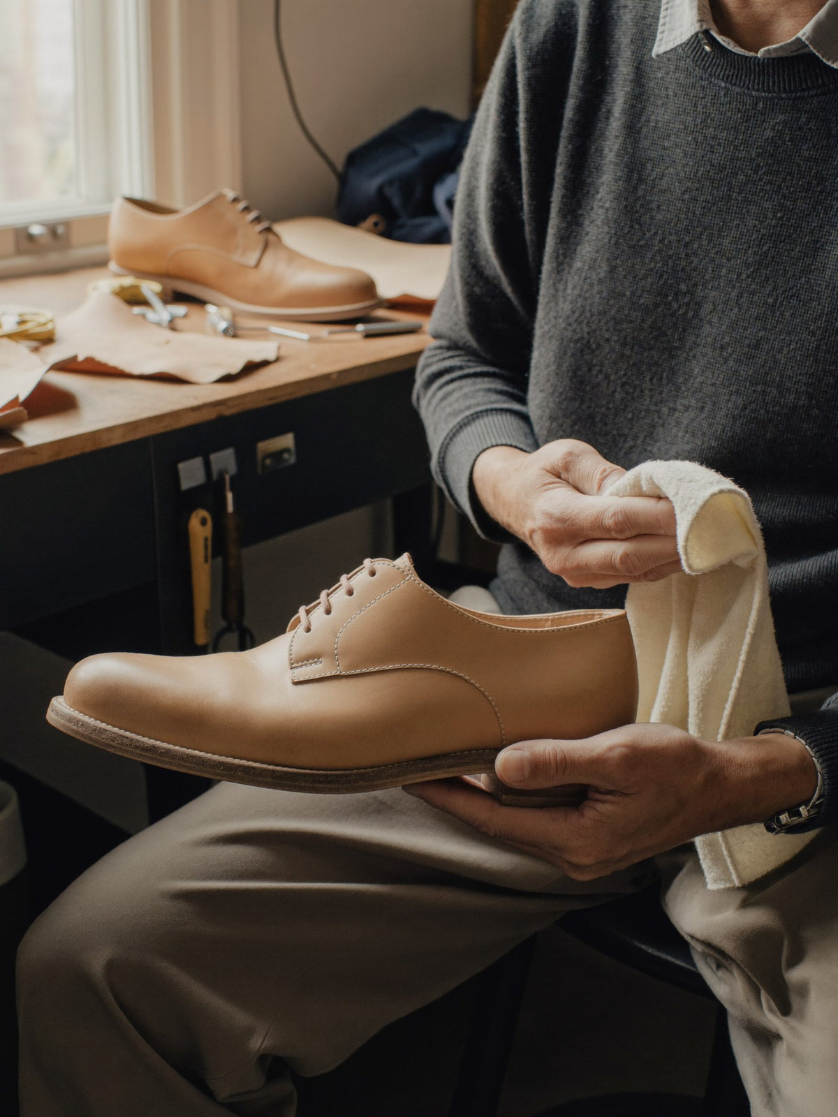 A shoemaker at a workbench buffing a tan leather shoe by hand